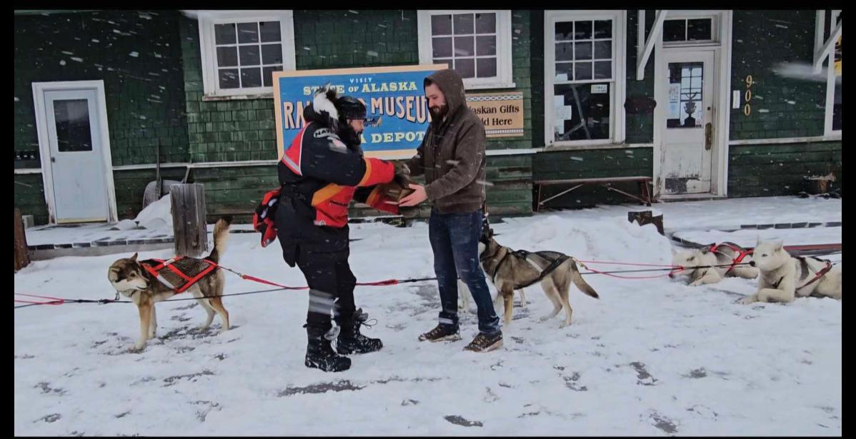 Photo by Adrienne Coffey EN ROUTE TO NOME–Nenana’s Mayor Joshua Verhagen hands off the box with the diphtheria antitoxin to Jonathan Hayes in a reenactment of the serum run that began January 27 in front of the Nenana railroad depot.